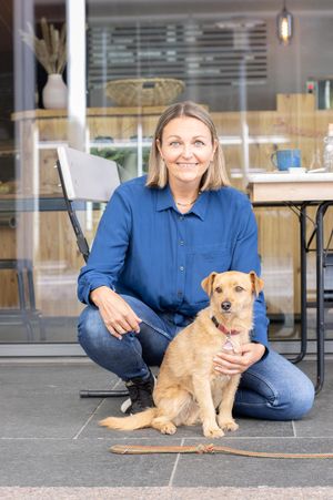 hellbrauner Terrier-Mix Hund sitzt vor hockender Frau, die ein blaues Hemd und Jeans trägt. Im Hintergrund sieht man einen Tisch und Stuhl vor einem Cafe