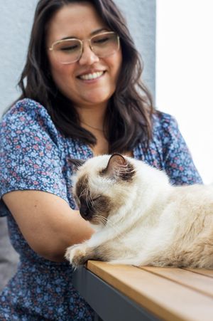 Frau in blauem Kleid sitzt an einem Holztisch und betrachtet lächelnd eine auf dem Tisch liegende Ragdoll Katze