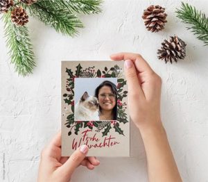 Hände halten eine Weihnachtskarte mit dem Foto einer Ragdoll-Katze zusammen mit einer Frau. Um die Hände herum liegen Tannenzweige und Zapfen