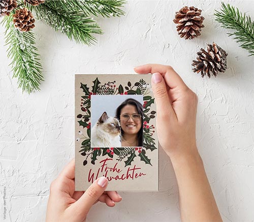 Hände halten eine Weihnachtskarte mit dem Foto einer Ragdoll-Katze zusammen mit einer Frau. Um die Hände herum liegen Tannenzweige und Zapfen